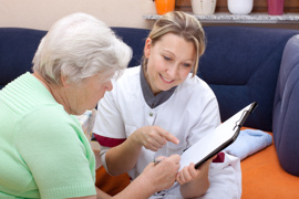Dame in Kleidung einer Krankenschwester sitzt neben älterer Dame und erklärt ein Formular