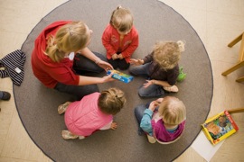 Erzieherin und Kindergartenkinder knien auf einem runden Teppich (Vogelperspektive)