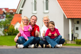 Erwachsene (Vater und Mutter) sitzen auf der Straße vor einem Haus (Eigenheim), jeweils ein Mädchen (Tochter) auf dem Schoß.